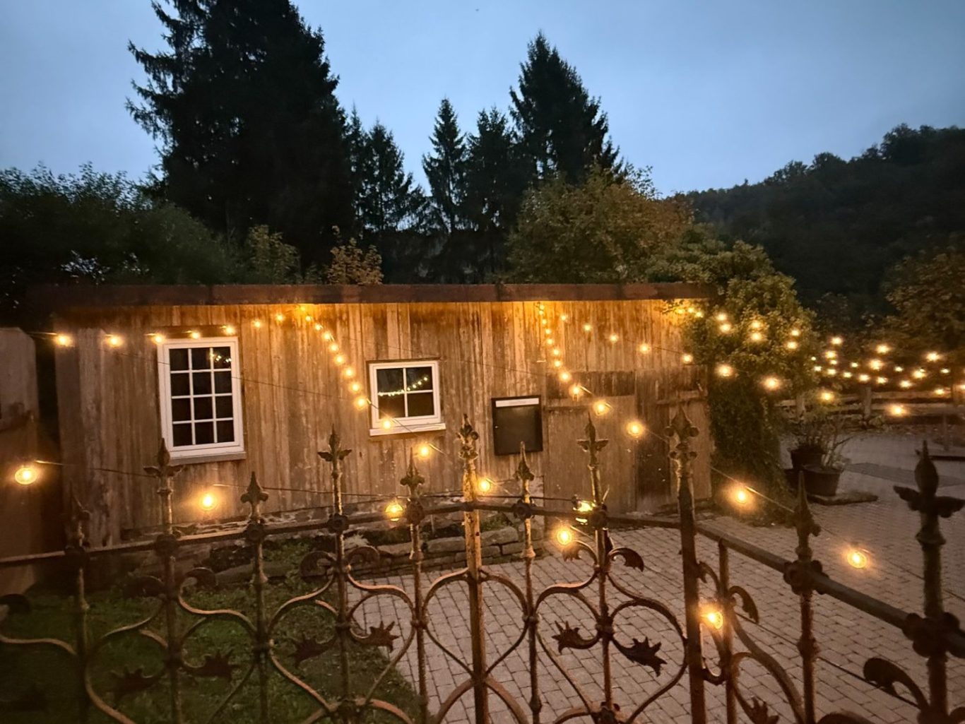 Blick auf die hölzerne Partyscheune von außen in der Dämmerung. Mit Hoffläche, Lichterkette, rustikalen Zaun. Natur im Hintergrund 